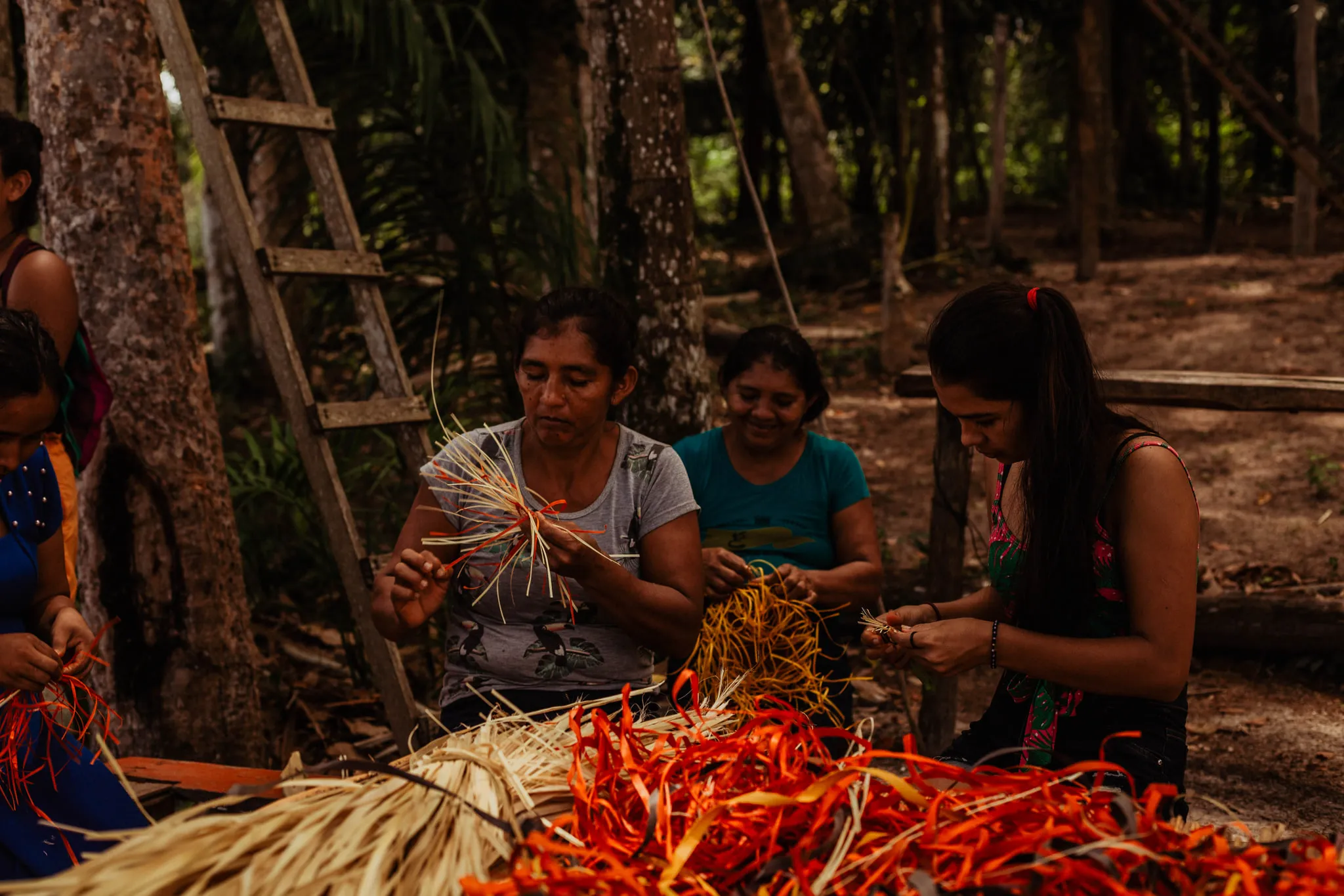 Five women from the community sitting on the ground among the trees, braiding bundles of straw dyed orange and red