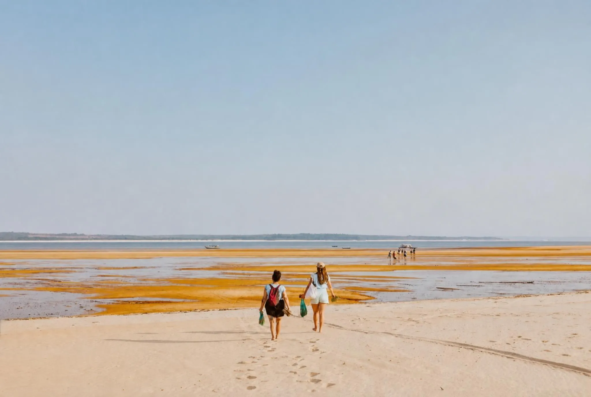 Two people walking away across an enormous pale sandbank, with more figures and moored boats far off at the waterline under a hazy sky