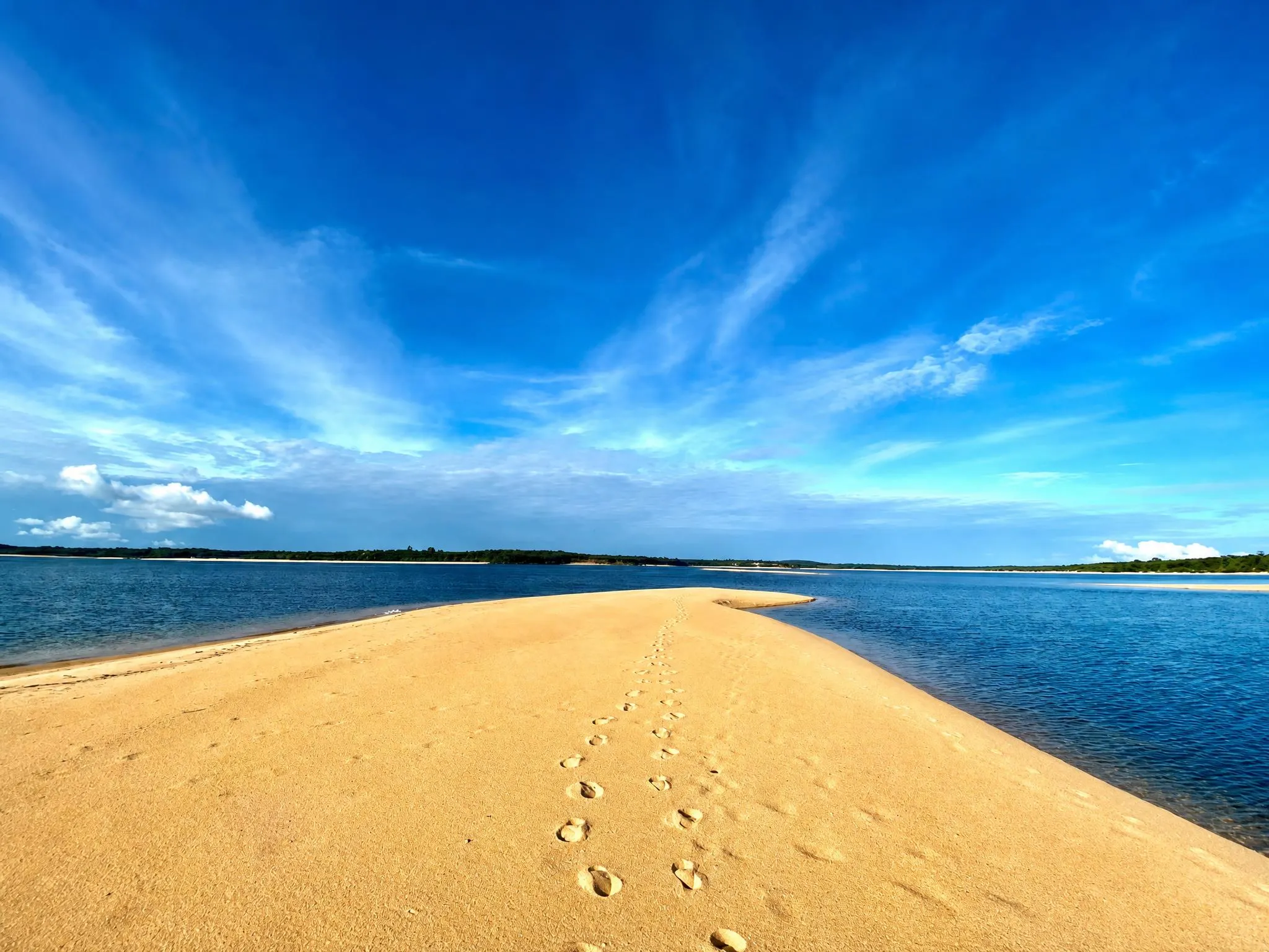 Golden sandbank stretching into the blue river, footprints in the sand and the tree line on the horizon