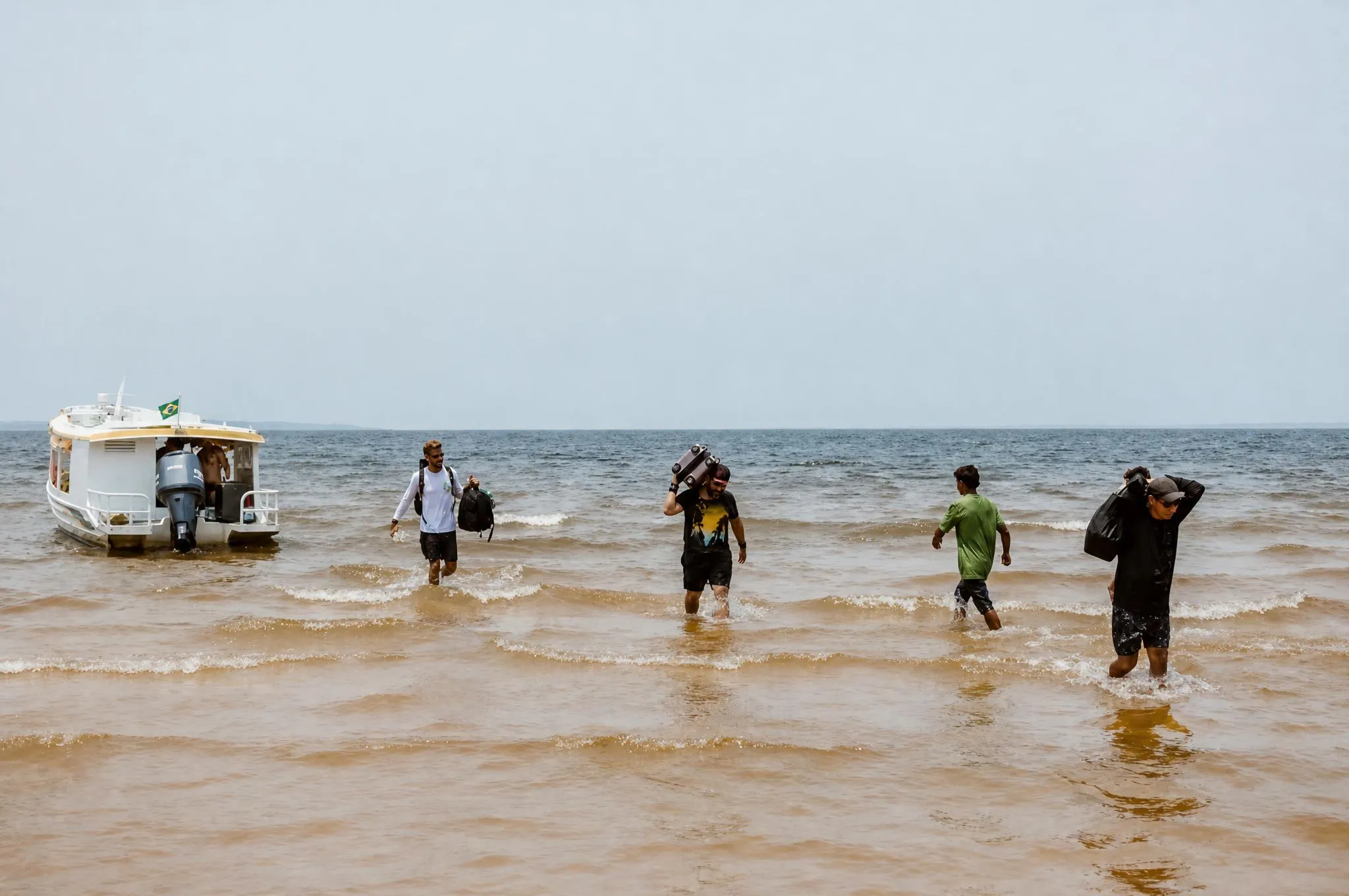 Pessoas desembarcando de um barco na água rasa da praia, com bagagem na mão