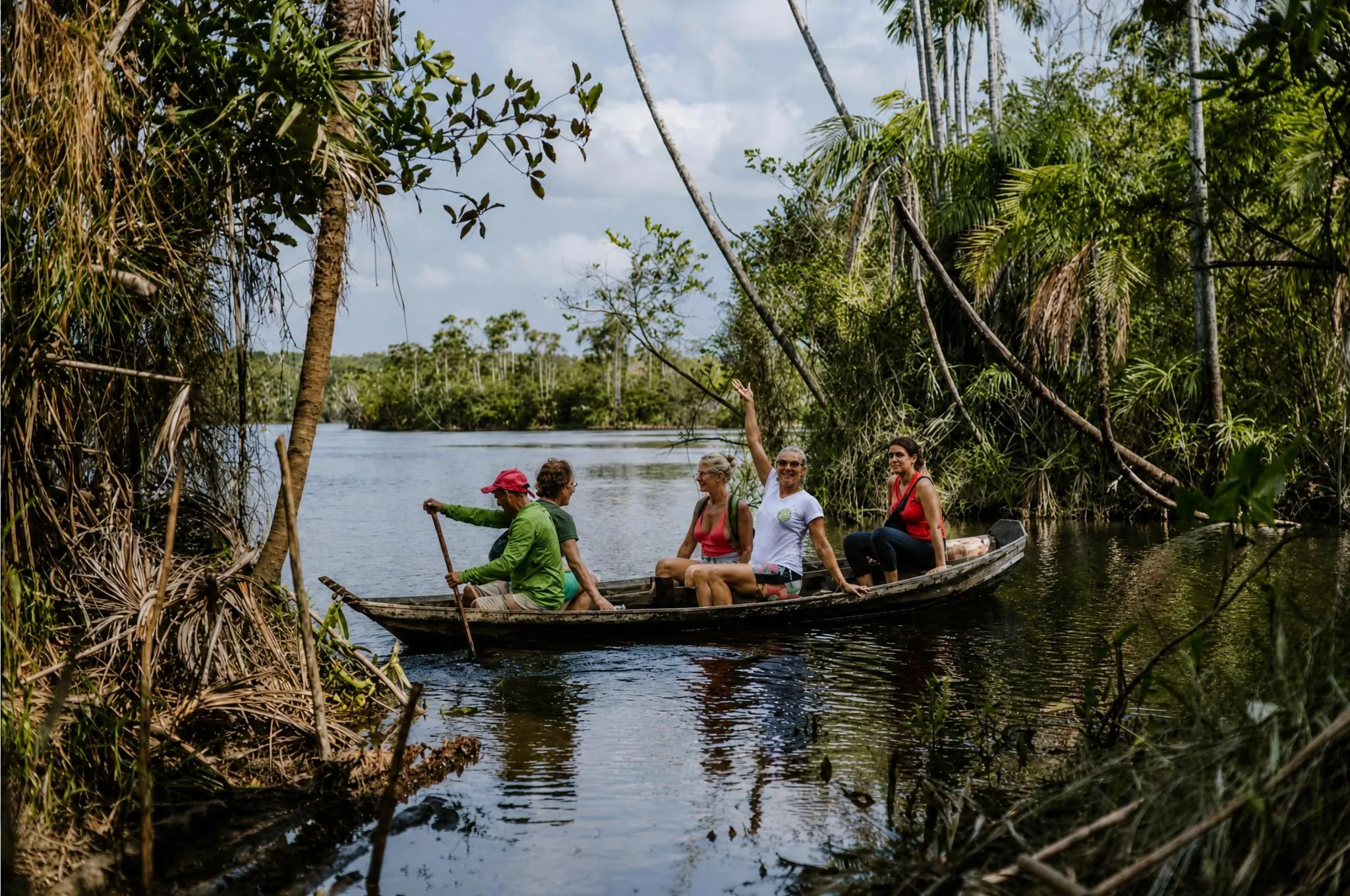 Four people inside a wooden canoe in the flooded forest, among submerged trunks and vines, on dark still water