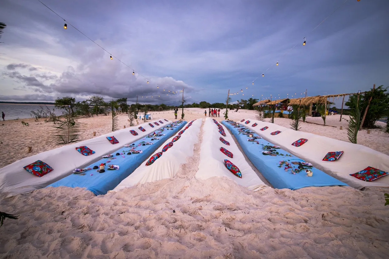 Long low tables set on the sand at dusk, dressed in blue and white, with floral cotton cushions and a strung line of lit bulbs overhead