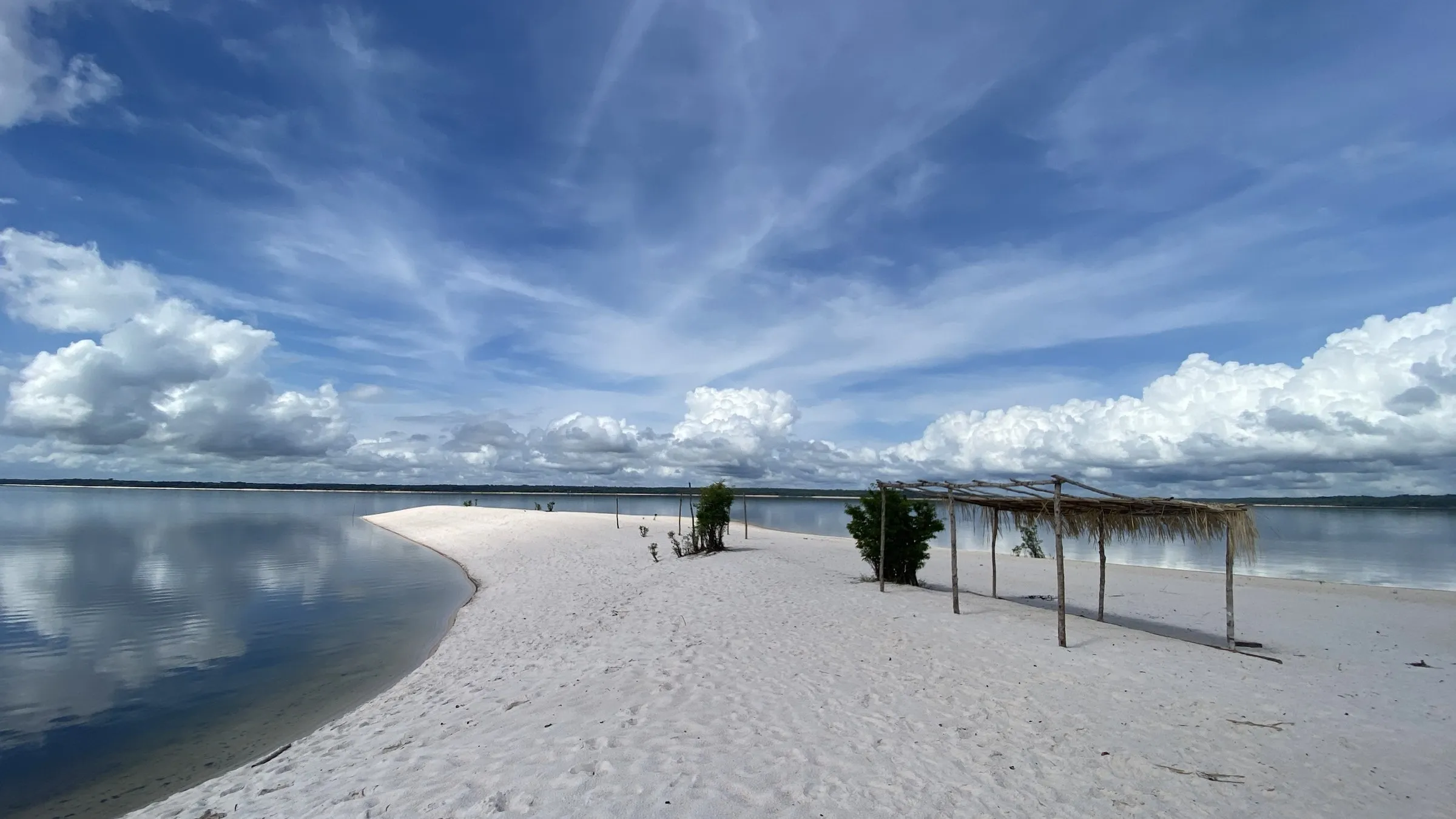 A white sandbank on the Rio Arapiuns, the still water mirroring a sky of high cloud, with a palm-thatched shade at the tip