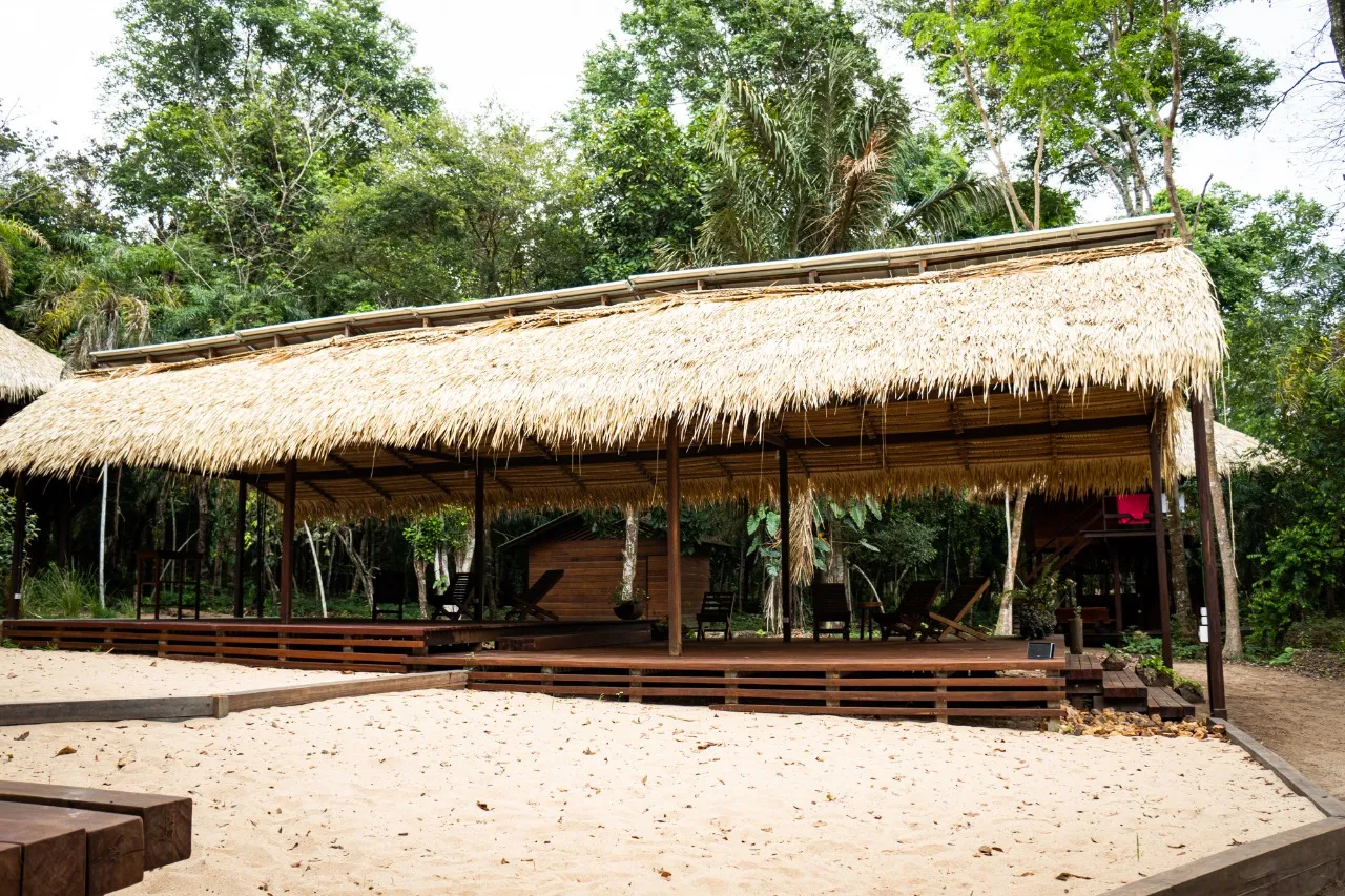 Long palm-thatched pavilion seen from the sand, with a timber deck, chairs and broad steps down to the pale ground