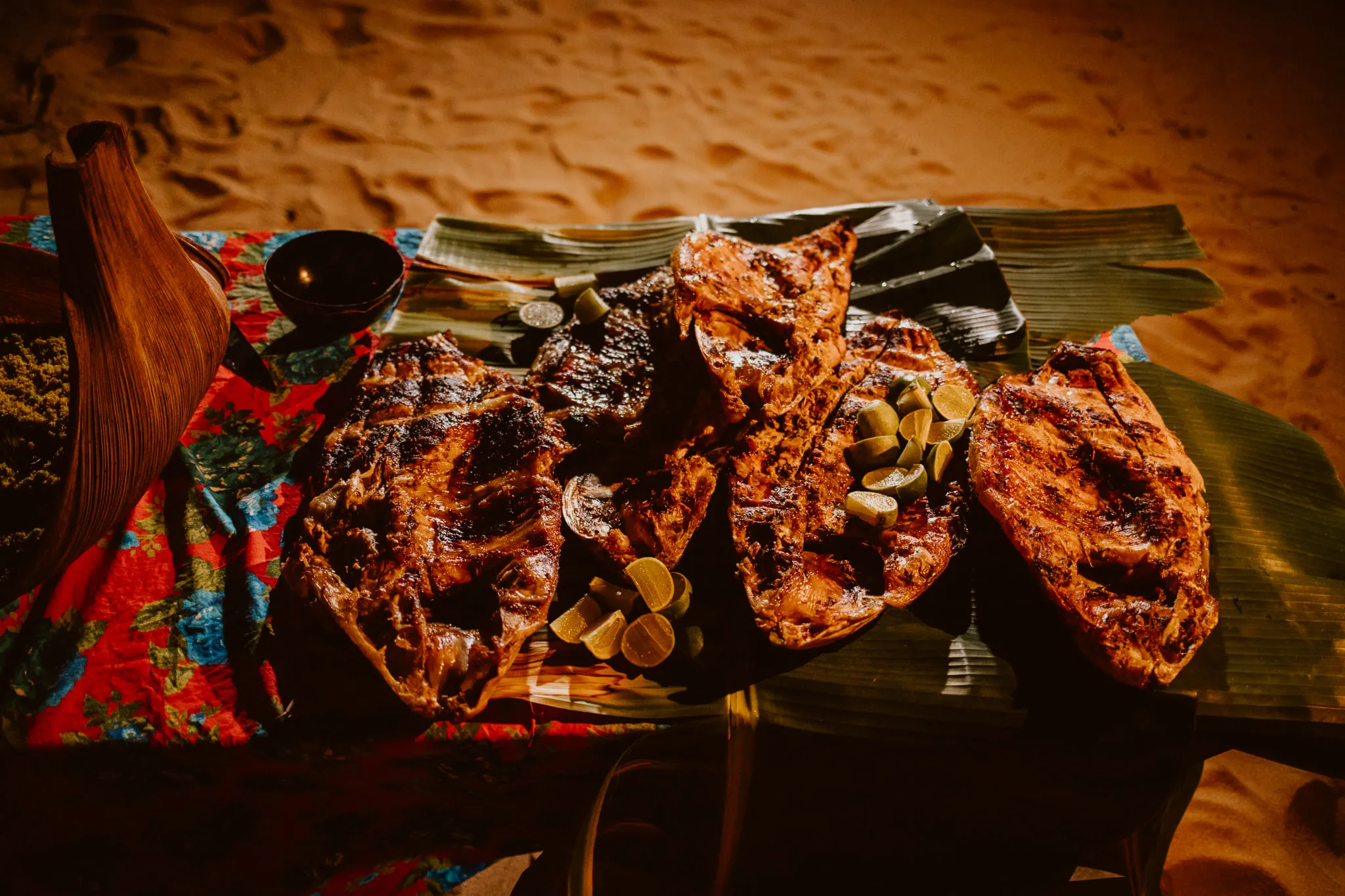 Large fish split and grilled over coals on banana leaves, with lime wedges, laid on a mat on the sand by candlelight