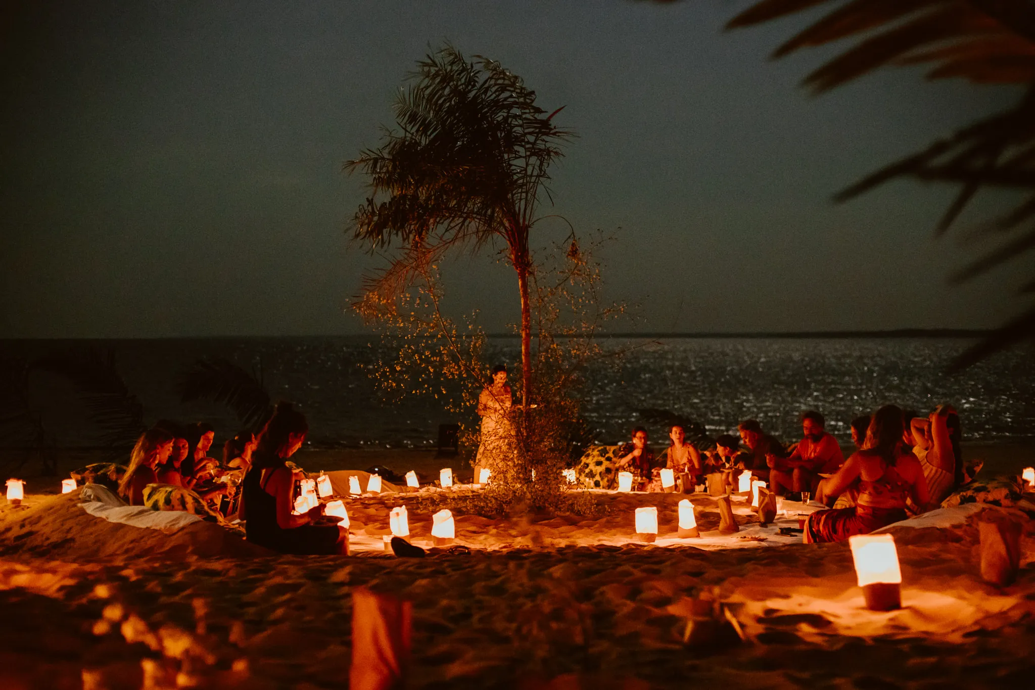 Large group seated in a circle on the sand at dusk, surrounded by lit lanterns, with a palm tree at the center and the river behind