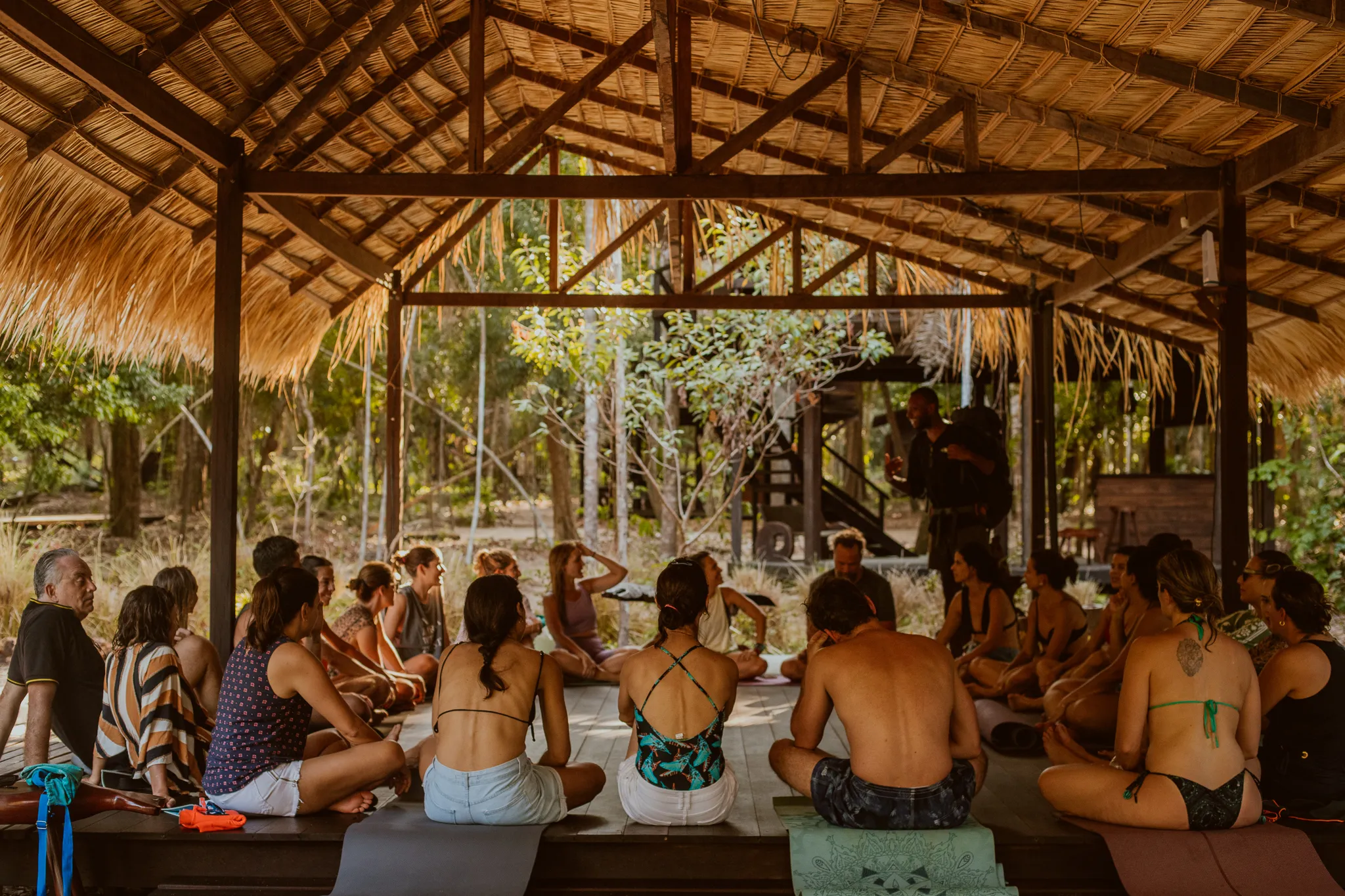 A circle of about twenty people sitting on mats on the wooden floor of the thatched pavilion, facing a facilitator who is standing, with the forest visible through the open bays