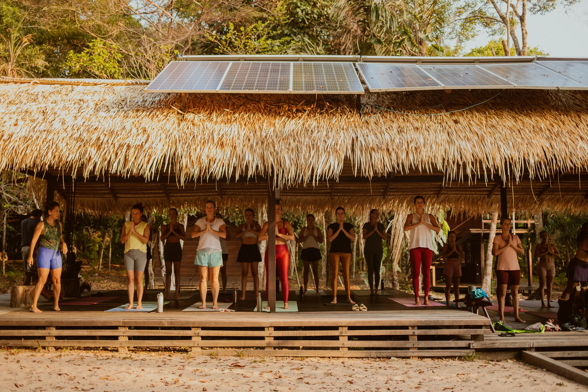 A dozen people standing on mats along a wooden deck under a long palm-thatched roof, hands together at the chest, with solar panels mounted above the roofline